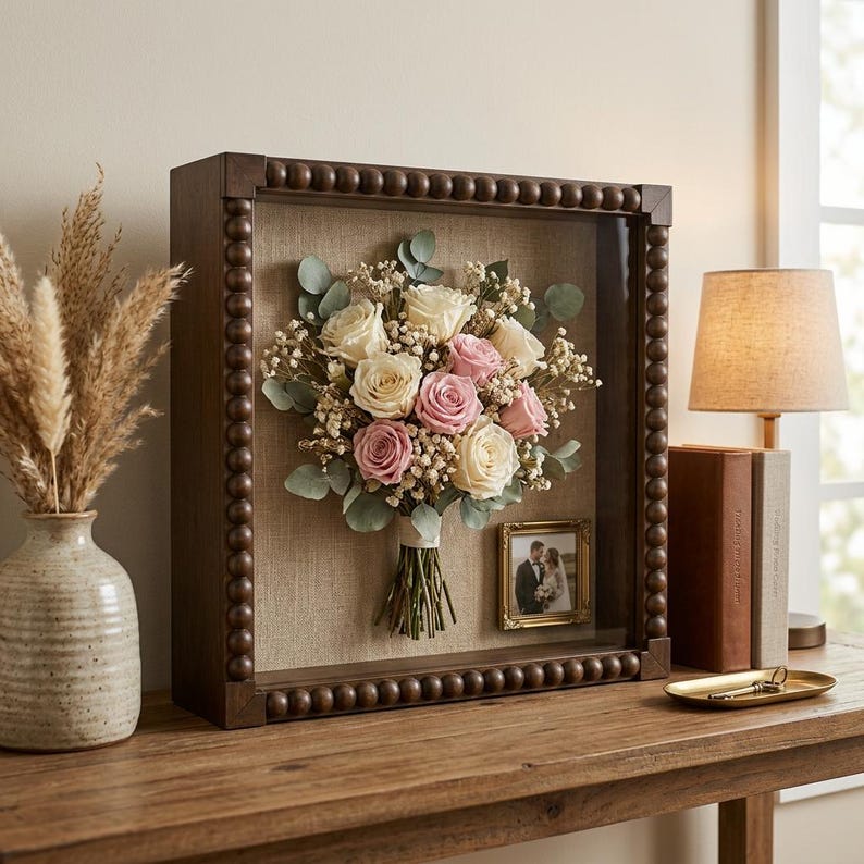 May include: A square shadow box frame with a dark brown beaded border containing a preserved bouquet of white and pink roses, baby's breath, and eucalyptus leaves. A small gold-toned picture frame holding a photograph is mounted inside.