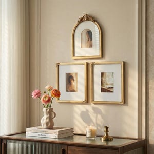 May include: Three gold-framed pictures of different shapes hang on a cream wall. A vase of pink and orange flowers sits on a wooden cabinet with books and a candle. The frames have decorative detailing.