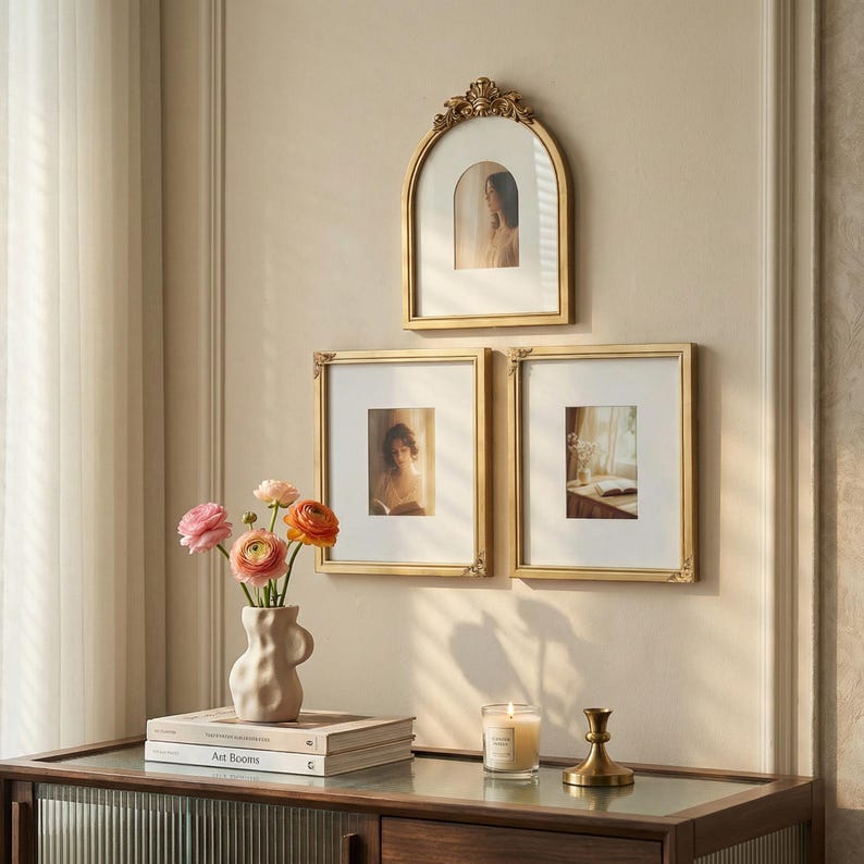 Puede incluir: Tres cuadros dorados de diferentes formas cuelgan de una pared beige. Un jarr&oacute;n con flores rosas y naranjas se encuentra sobre un mueble de madera con libros y una vela. Los marcos tienen detalles ornamentados.