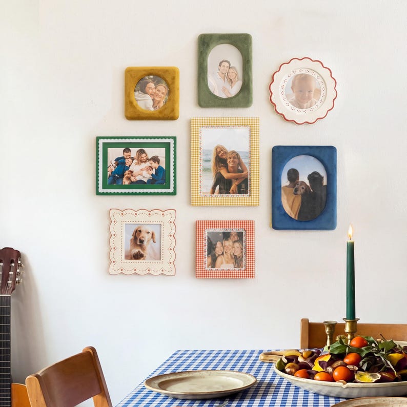 May include: A collection of eight wall-mounted picture frames in various shapes, colors, and textures, including velvet, scalloped edges, and gingham patterns, displaying photographs of people and a dog, arranged on a plain white wall.