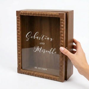 Puede incluir: Una caja de sombra de madera con frente de vidrio y puerta con bisagras. La caja est&aacute; grabada con los nombres "Sebastian y Mireille" y la fecha "05.25.2024". El marco tiene un borde decorativo con cuentas.