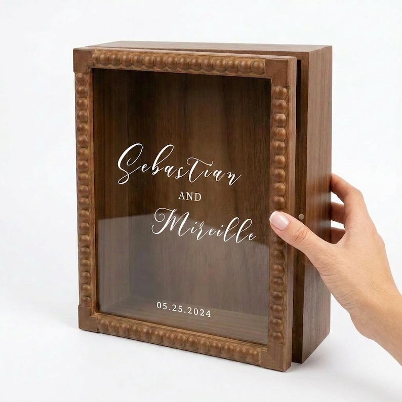 Puede incluir: Una caja de sombra de madera con frente de vidrio y puerta con bisagras. La caja est&aacute; grabada con los nombres "Sebastian y Mireille" y la fecha "05.25.2024". El marco tiene un borde decorativo con cuentas.