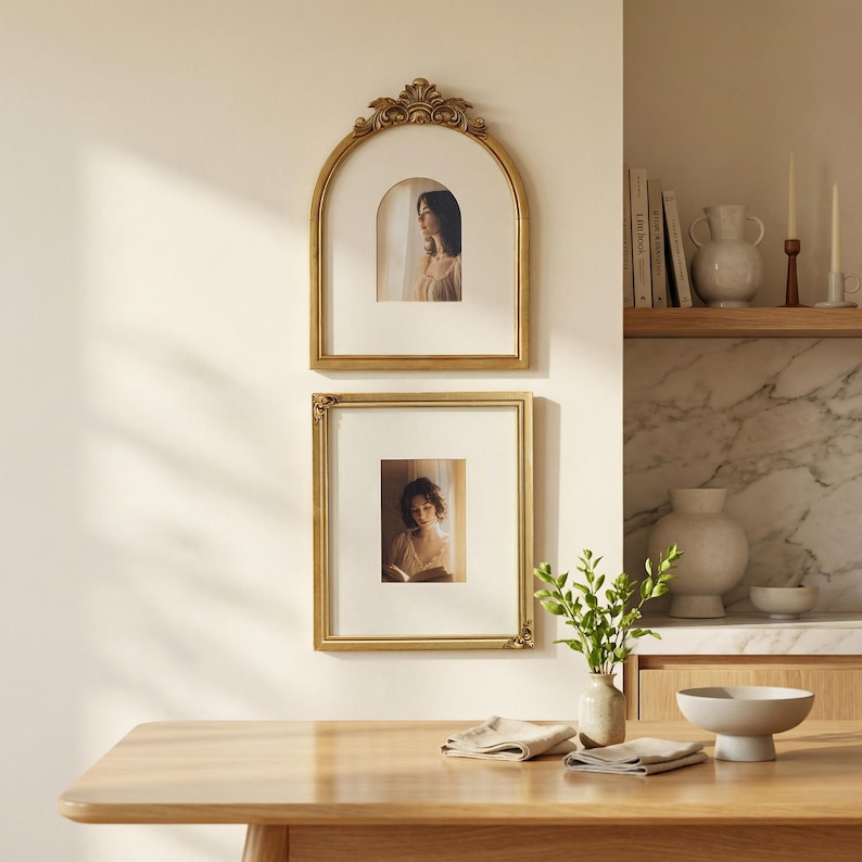 Puede incluir: Dos cuadros con marcos dorados cuelgan de una pared color crema. El marco superior es arqueado con detalles ornamentados, y el marco inferior es rectangular. Ambos marcos contienen retratos de mujeres. Una mesa de madera est&aacute; en primer plano.