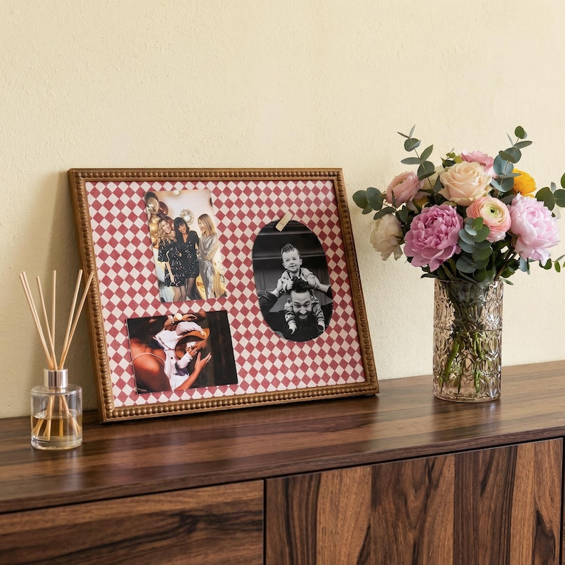 May include: A framed photo display with a red and white diamond pattern holds three photos. The frame is a warm brown color. A glass vase with a bouquet of pink, yellow, and white flowers sits to the right. A reed diffuser is on the left.
