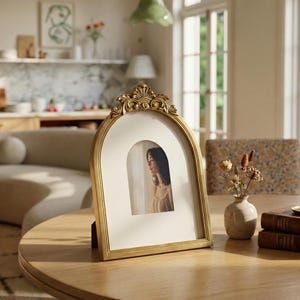 Puede incluir: Un marco de fotos dorado en forma de arco con detalles ornamentados. El marco contiene una fotograf&iacute;a de una persona mirando por una ventana. Se exhibe sobre una mesa de madera, con un peque&ntilde;o jarr&oacute;n de flores secas y libros cerca.