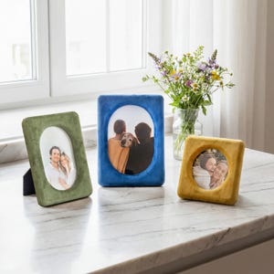 May include: Three velvet-covered picture frames in olive green, royal blue, and mustard yellow. The olive frame is rectangular with an oval opening, the blue frame is rectangular with an oval opening, and the yellow frame is square with a circular opening.