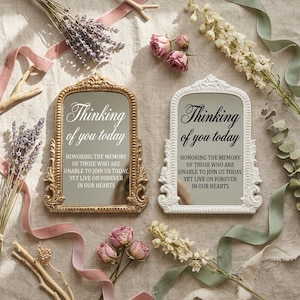 Könnte beinhalten: Zwei dekorative Spiegel mit verzierten Rahmen, einer goldfarben und einer weiß, jeweils mit der Aufschrift "Thinking of you today". Die Spiegel sind von Trockenblumen, Bändern und Rosen umgeben, was ein Gedenkthema erzeugt.