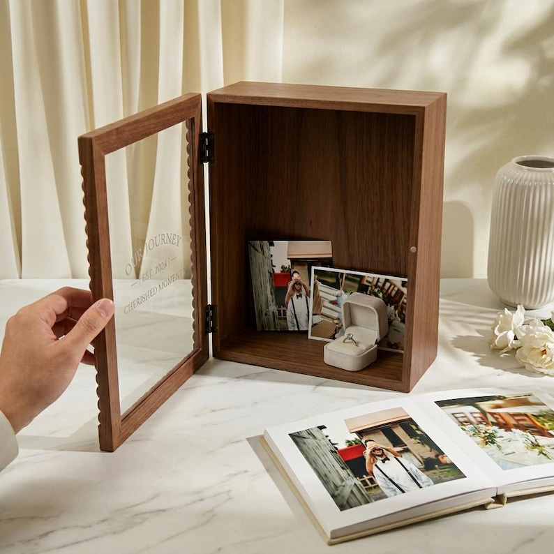 Puede incluir: Una caja de exhibici&oacute;n de madera con puerta de cristal, que contiene fotograf&iacute;as y un joyero. La puerta tiene el texto "Our Journey" y "Cherished Moments". La caja est&aacute; abierta, revelando su contenido. Un &aacute;lbum de fotos abierto est&aacute; en primer plano.