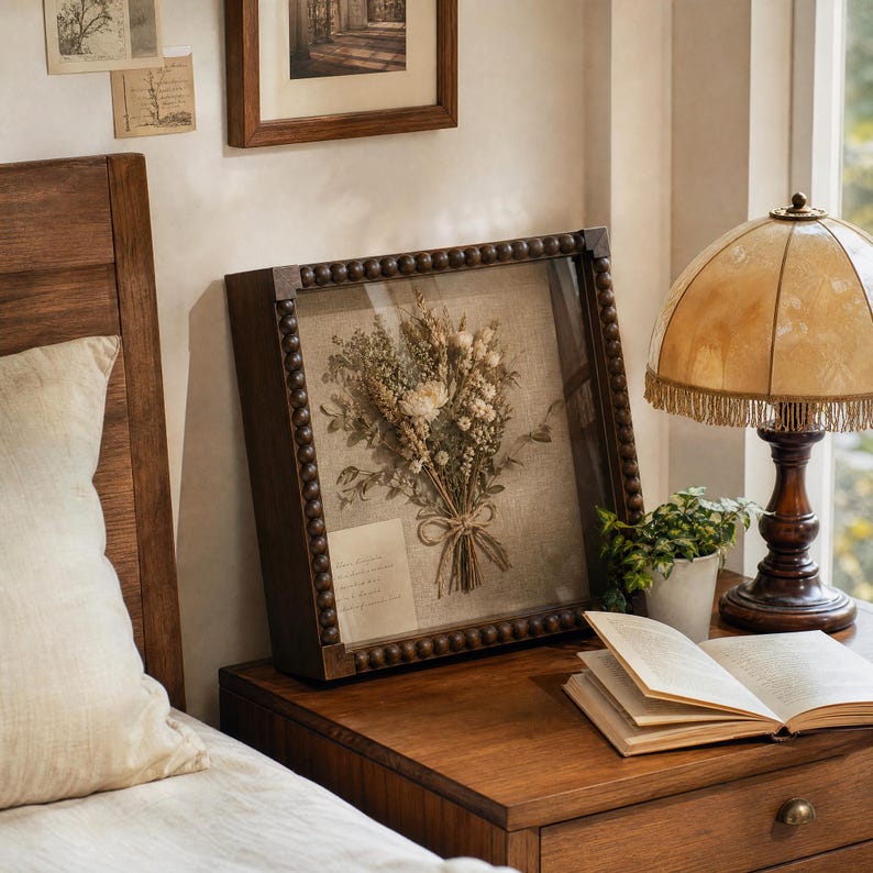 May include: A square wooden shadow box frame with a beaded border containing a dried flower bouquet arranged on a light beige linen background. The frame features a glass front and a small rectangular paper note tucked into the bottom left corner.