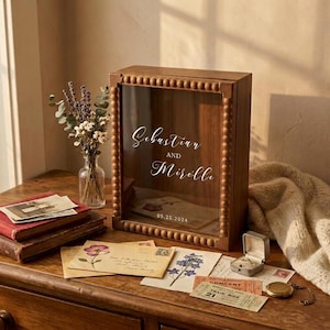 Puede incluir: Una caja expositora de madera con frente de cristal, con los nombres "Sebastian y Mirelle" y la fecha "05.25.2024". La caja est&aacute; rodeada de cuentas de madera decorativas. Cartas antiguas, un joyero y un reloj de bolsillo est&aacute;n sobre una superficie de madera.