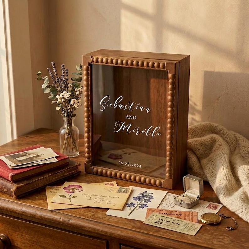 Puede incluir: Una caja expositora de madera con frente de cristal, con los nombres "Sebastian y Mirelle" y la fecha "05.25.2024". La caja est&aacute; rodeada de cuentas de madera decorativas. Cartas antiguas, un joyero y un reloj de bolsillo est&aacute;n sobre una superficie de madera.