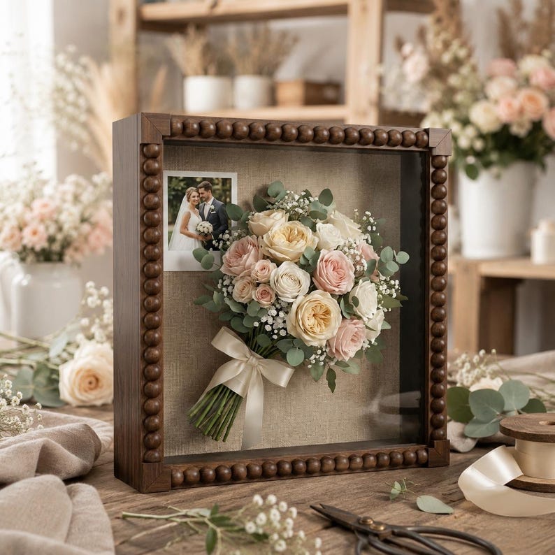 May include: A square shadow box frame with a dark wood finish and beaded edges, containing a preserved wedding bouquet of cream and pink roses with eucalyptus and baby's breath, tied with a cream ribbon, next to a small square wedding photograph.