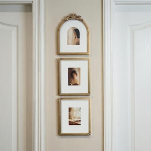 Puede incluir: Tres marcos dorados colgados en una pared beige. El marco superior es arqueado, los otros dos rectangulares. Cada marco contiene una fotograf&iacute;a con un paspart&uacute; blanco. Los marcos tienen detalles ornamentados.