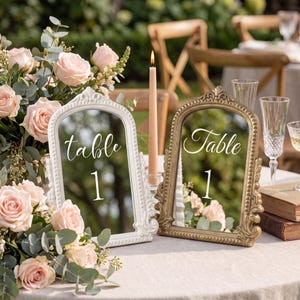 Könnte beinhalten: Zwei gewölbte Tischnummernschilder mit verzierten Rahmen, eines in Weiß und eines in Gold, die jeweils den weißen Text "table 1" zeigen. Die Schilder stehen auf einem Tisch neben einer hohen Stabkerze, einem Strauß blassrosa Rosen und Gläsern.