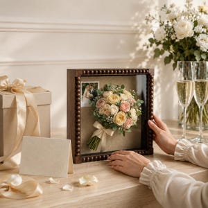 May include: A square wooden shadow box frame with a beaded border containing a preserved bouquet of cream and pink roses, greenery, and a small photograph of a couple. The frame sits on a table next to a gift box with a ribbon and a blank card.