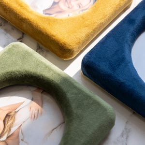 May include: Three square picture frames with rounded corners, each covered in a soft, plush velvet fabric. The frames are colored in mustard yellow, deep navy blue, and sage green, and each holds a photograph of a person.