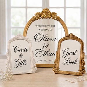 May include: Three arched signs with ornate frames. The large central gold sign reads: WELCOME TO THE WEDDING OF Olivia & Ethan JUNE 21, 2027. The left white sign reads: Cards & Gifts. The right gold sign reads: Guest Book.
