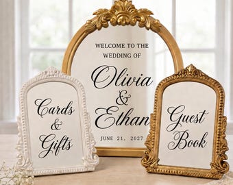 May include: Three arched signs with ornate frames. The large central gold sign reads: WELCOME TO THE WEDDING OF Olivia & Ethan JUNE 21, 2027. The left white sign reads: Cards & Gifts. The right gold sign reads: Guest Book.