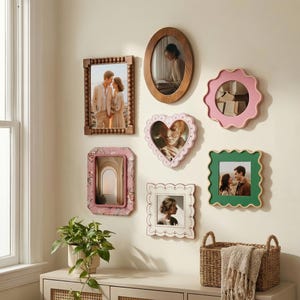 Può includere: Una collezione di cornici decorative per foto in varie forme e colori, tra cui design a cuore, ovali e smerlati. Le cornici sono montate su una parete color crema, che mostra fotografie. Sono visibili anche un cesto di vimini e una pianta in vaso.