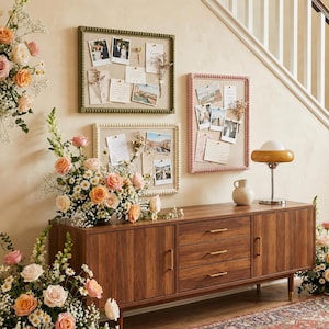 Puede incluir: Tres tableros de anuncios decorativos con fotos y notas cuelgan de una pared sobre un mueble de madera. Los tableros tienen marcos de diferentes colores: verde, rosa y blanco. Una lámpara y un jarrón están sobre el mueble, y hay flores en la habitación.
