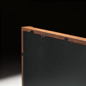 May include: Close-up of the back of a rectangular wooden picture frame. The frame has a dark brown finish and a black backing. Metal hanging hardware is attached to the frame.