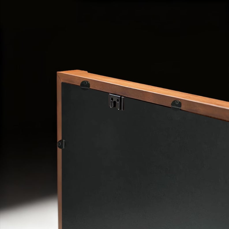 May include: Close-up of the back of a rectangular wooden picture frame. The frame has a dark brown finish and a black backing. Metal hanging hardware is attached to the frame.