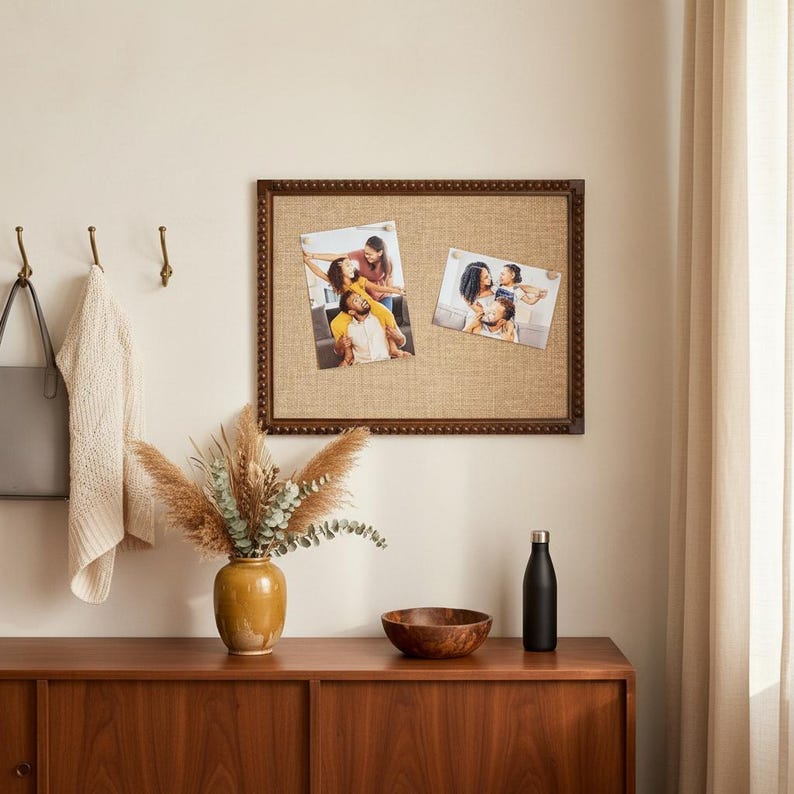 May include: A framed noticeboard with a hessian surface displays two photographs. A wooden cabinet holds a vase with dried flowers, a wooden bowl, and a black water bottle. A coat and bag hang on hooks.