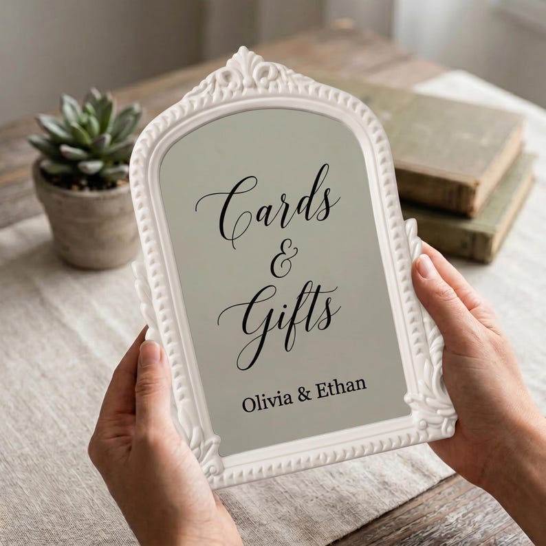 May include: An arched white ornate frame containing a light gray sign with black text that reads Cards & Gifts and Olivia & Ethan. The frame features decorative scrollwork along the edges and is held in a vertical orientation against a neutral background.