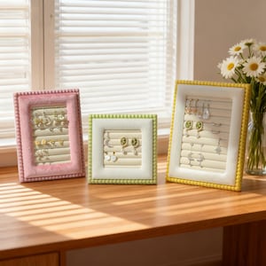 May include: Three earring display frames in pink, green, and yellow. Each frame has a plush border and a cream-colored interior with slots for hanging earrings. The frames are arranged on a wooden surface near a window with daisies.