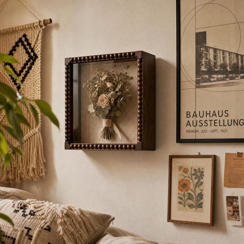 May include: A square shadow box frame with a dark brown wooden finish and a beaded border detail. Inside the glass-fronted frame, a dried floral bouquet featuring light-colored roses and greenery is mounted against a neutral linen backing.
