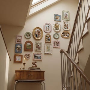 Puede incluir: Una colección de fotografías enmarcadas de diversas formas y tamaños adorna una pared. Los marcos son de color dorado, azul, rosa y blanco, creando una exhibición estilo galería. Debajo, un mueble de madera con un candelabro y objetos decorativos.