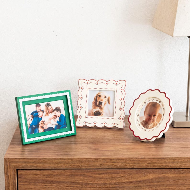 May include: Three decorative picture frames displayed together. One is a rectangular green frame with a white scalloped border. The second is a square cream frame with a red scalloped edge and heart details. The third is a round cream frame with a red scalloped edge.