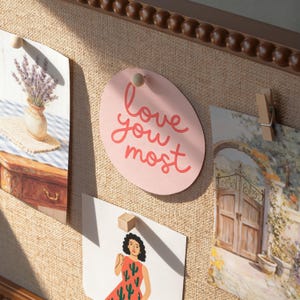 May include: A noticeboard displaying several images, including a pink oval card with the words "love you most" in red. Other images include a vase of lavender, a woman in a dress, and a landscape scene.