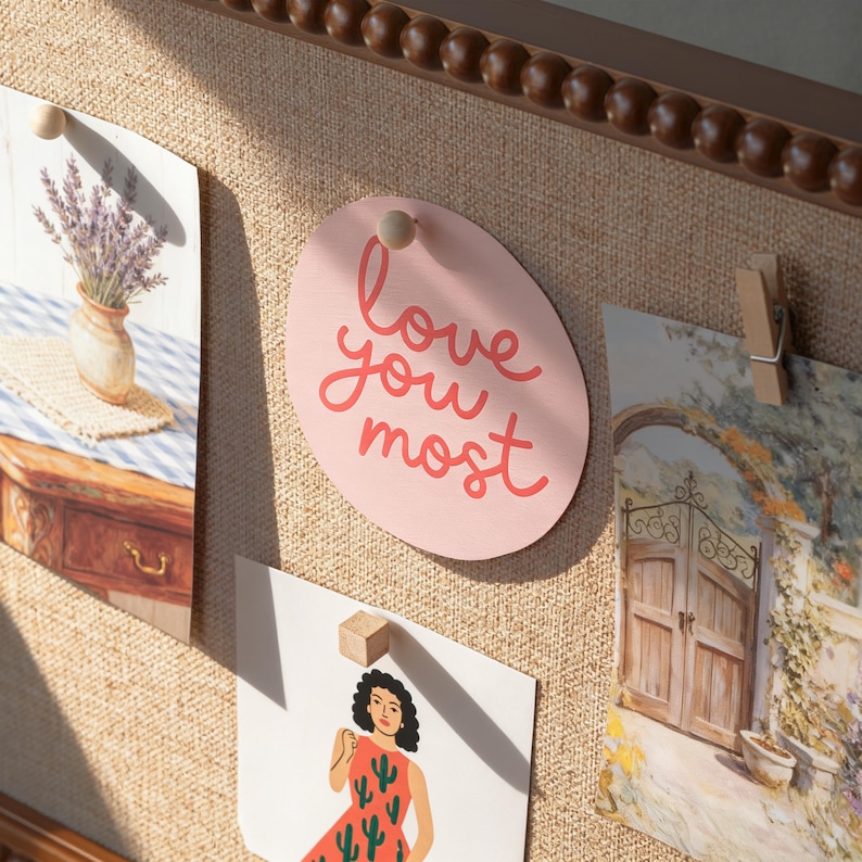 May include: A noticeboard displaying several images, including a pink oval card with the words "love you most" in red. Other images include a vase of lavender, a woman in a dress, and a landscape scene.