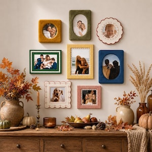 Op de afbeelding: Een galerijwand met negen verschillende fotolijsten in diverse vormen, kleuren en texturen, waaronder fluweel, gingham en geschulpte randen, met persoonlijke foto's, opgehangen boven een houten sidetable.