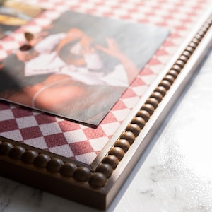 May include: A rectangular wooden picture frame with a red and white checkered fabric background. The frame has a dark brown beaded trim and holds a photograph. The photo is slightly out of focus, showing a person.