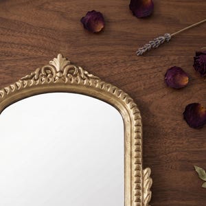 May include: An arched wall mirror featuring a metallic gold frame with ornate, raised scrollwork detailing at the top and a textured, repeating pattern along the sides. The mirror has a reflective glass surface and a distinct, decorative crest.