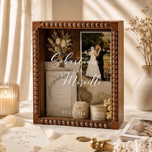 Könnte beinhalten: Eine Holz-Schattenbox mit Perlenrand zeigt ein Hochzeitsfoto, eine Einladung, einen Ring und eine Kerze. Das Foto zeigt ein sich küssendes Paar. Auf der Einladung steht "Sebastian und Mireille" mit dem Datum "05.25.2024". Trockenblumen und eine Kerze sind ebenfalls enthalten.