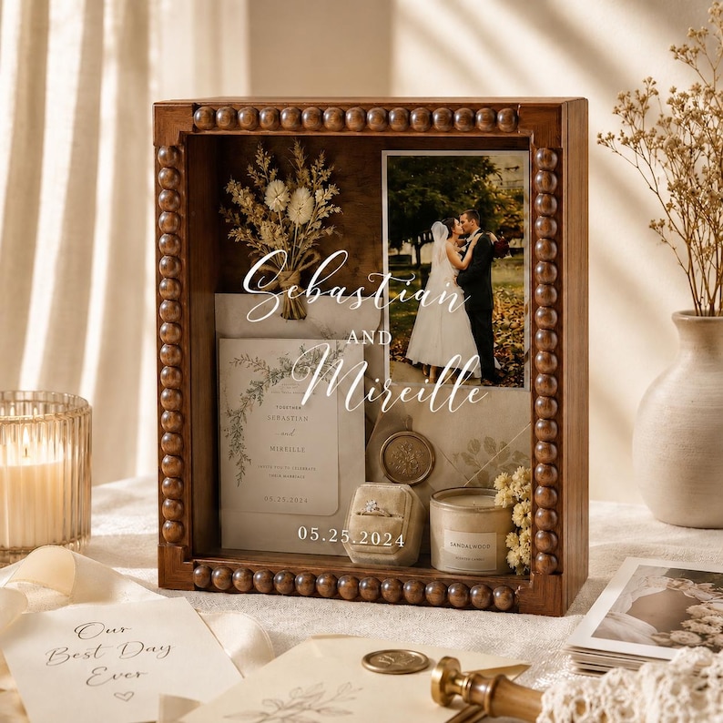 Puede incluir: Una caja de sombra de madera con borde de cuentas muestra una foto de boda, invitaci&oacute;n, anillo y vela. La foto muestra a una pareja bes&aacute;ndose. La invitaci&oacute;n dice "Sebastian y Mireille" con la fecha "05.25.2024". Flores secas y una vela tambi&eacute;n est&aacute;n dentro.