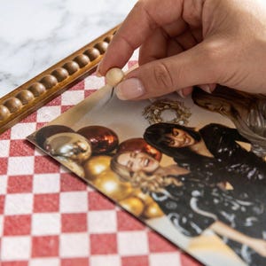 May include: A wooden frame with a decorative border holds a photograph of three women. A hand is shown placing a small, round, wooden push pin into the photograph. The photograph is on a red and white checkered surface.