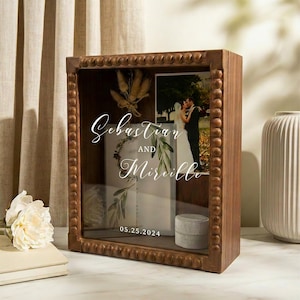 Puede incluir: Una caja de sombra de madera con frente de vidrio, que muestra una foto de boda y una invitaci&oacute;n. La caja est&aacute; adornada con cuentas decorativas y presenta los nombres "Sebastian and Mireille" y la fecha "05.25.2024" en escritura elegante.