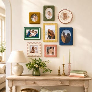 May include: A gallery wall arrangement featuring eight assorted picture frames in various shapes, colors, and textures, including velvet, scalloped, and patterned designs, displayed above a wooden console table with a lamp and decor.