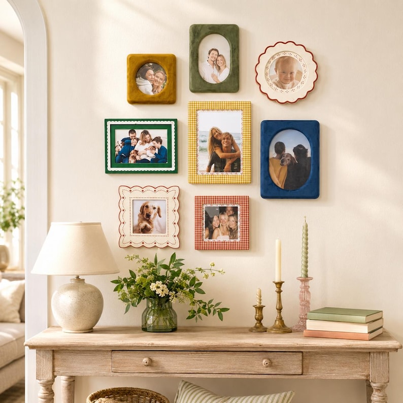 May include: A gallery wall arrangement featuring eight assorted picture frames in various shapes, colors, and textures, including velvet, scalloped, and patterned designs, displayed above a wooden console table with a lamp and decor.