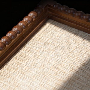 May include: Close-up of a wooden picture frame with a light tan linen backing. The frame is a dark brown colour and has a decorative beaded edge. The background is black.