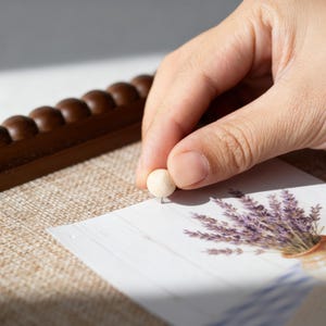 May include: A small, light-coloured wooden drawing pin being pressed into a card featuring a lavender bouquet illustration. The card rests on a textured surface, with a dark brown wooden element behind.