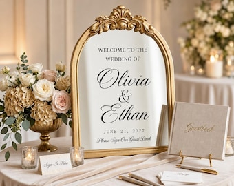May include: An arched gold-framed mirror sign with black text reading Welcome to the Wedding of Olivia & Ethan June 21, 2027 Please Sign Our Guest Book. Includes a beige guestbook, a Sign-In Here card, pens, and a floral arrangement in a gold vase.