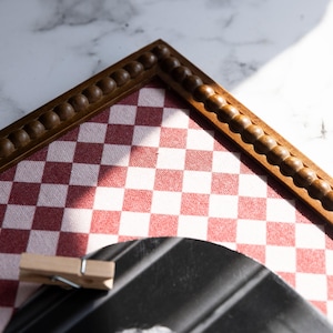 May include: A wooden framed board with a red and white checkered pattern. The frame has a decorative beaded edge. A wooden clothespin is clipped to a black object in the foreground.
