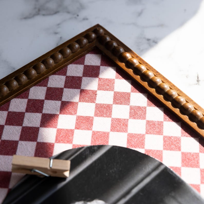 May include: A wooden framed board with a red and white checkered pattern. The frame has a decorative beaded edge. A wooden clothespin is clipped to a black object in the foreground.