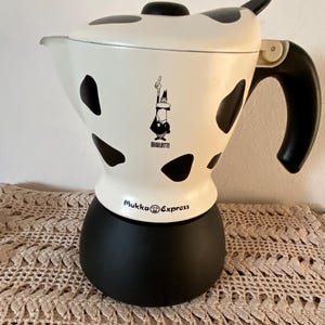 Peut inclure: Une cafetière Bialetti Mukka Express blanche et noire. La cafetière a une base et une poignée noires, avec des taches noires sur la partie supérieure blanche. Le logo Bialetti est visible à l'avant.