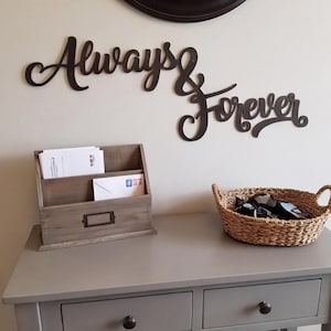 Può includere: Una scritta da parete marrone scuro con il testo in corsivo Always & Forever. Accanto si trova un portaposta in legno a due scomparti contenente buste bianche e un cesto in erba marina intrecciata con un paio di scarpe bianche e nere.